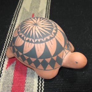 May include: A terracotta-colored ceramic turtle figurine with a detailed geometric design in dark gray on its shell. The turtle has a rounded body, a small head, and four stubby legs. The design features radiating lines and diamond shapes.