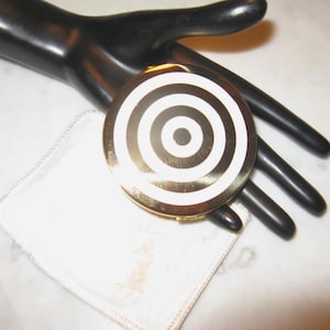 May include: A gold-toned compact with a concentric black and white design. The compact is round and sits on a white cloth. The background is a black hand and a white surface.