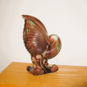 May include: A brown and green glazed ceramic seashell vase with a swirling design. The vase is shaped like a seashell with a curled tail and a base that resembles waves.