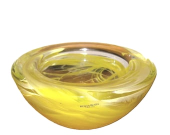 May include: A round glass bowl featuring swirling patterns of bright yellow and clear glass. The bowl has a thick, curved rim and a smooth, polished surface. The text KOSTA BODA is printed in small black letters on the side of the bowl.