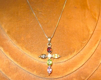 Puede incluir: Un collar de cadena de plata con un colgante de cruz con siete piedras preciosas ovaladas de varios colores, incluyendo rojo, blanco, amarillo, verde y morado.