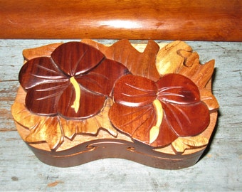 May include: A wooden puzzle box featuring a carved design of two hibiscus flowers. The box is crafted from various shades of brown wood and has a natural finish.