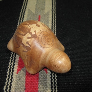 May include: A light brown, hand-carved ceramic sculpture with a rounded shape and a smooth finish. The sculpture features etched designs, including a spiral and a figure. The piece is displayed on a black and red patterned textile.