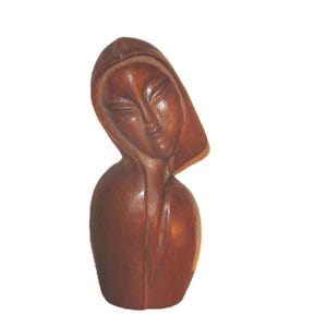 May include: A wooden sculpture of a woman's bust, featuring a serene expression and a hooded head covering. The sculpture is carved from a dark brown wood, with smooth, flowing lines and a polished finish. The bust is a decorative art piece.