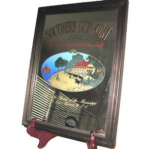 Puede incluir: Espejo enmarcado con las palabras "SOUTHERN COMFORT" en la parte superior. El espejo tiene un marco de madera marrón oscuro y presenta una imagen ovalada de una casa y un barco fluvial. También es visible el texto "The Grand Old Drink of the South".