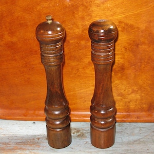 Może przedstawiać: Dwa drewniane solniczki i pieprzniczki z ciemnobrązowym wykończeniem. Solniczki mają zaokrąglony wierzchołek i wypukłe ciało.