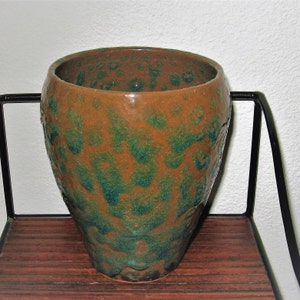 Puede incluir: Un jarrón de cerámica marrón y verde con una superficie texturizada. El jarrón tiene una boca ancha y una base estrecha.