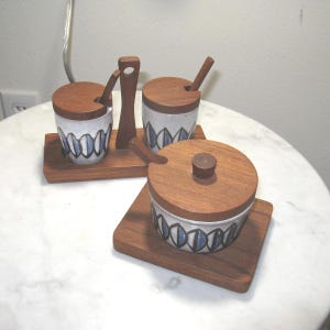 May include: A set of three ceramic and wood kitchen canisters. The canisters have wooden lids and are decorated with a blue and white geometric pattern. They sit on wooden trays, with a wooden spoon and a serving utensil.