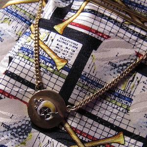 May include: A gold chain necklace with a round pendant featuring a stylized design. The necklace is lying on a fabric with a grid pattern and a repeating design of bones and the text "UNCLASSIFIED RATINGS SLOPE".