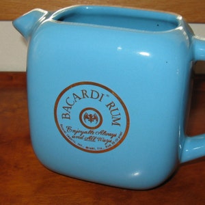 Può includere: Una brocca in ceramica azzurra di forma quadrata con un manico. La brocca ha un beccuccio e un logo circolare dorato che recita "BACARDI RUM" con il testo "Enjoyable Always and All Ways".