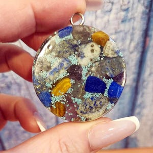 May include: A round pendant with a mix of blue, yellow, and gray stones set in clear resin. The pendant has a silver-colored bail at the top for attaching to a chain. The stones are arranged in a mosaic pattern.