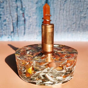 Könnte beinhalten: Ein dekorativer Kerzenhalter mit einer Basis aus klarem Harz, in die Metallflocken und getrocknete Pflanzen eingebettet sind. Ein kupferfarbenes Metallrohr erhebt sich von der Basis und trägt einen kleinen orangefarbenen Kristall mit einem spiralförmigen Metallakzent.