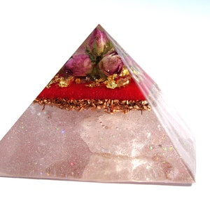 May include: A clear quartz pyramid with a red layer and gold flakes, topped with dried pink roses. The pyramid is filled with glitter and has a sparkly finish.