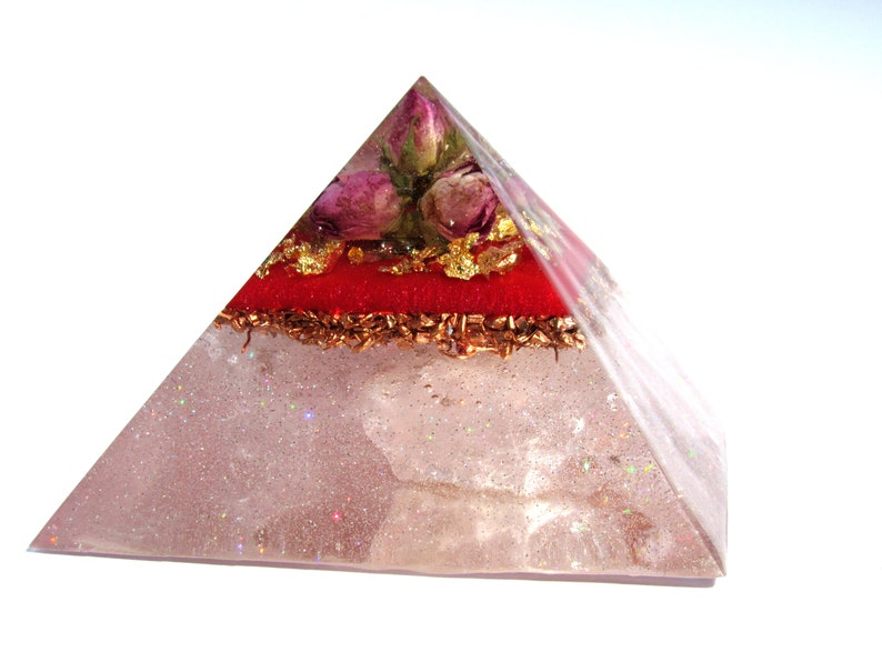 May include: A clear quartz pyramid with a red layer and gold flakes, topped with dried pink roses. The pyramid is filled with glitter and has a sparkly finish.