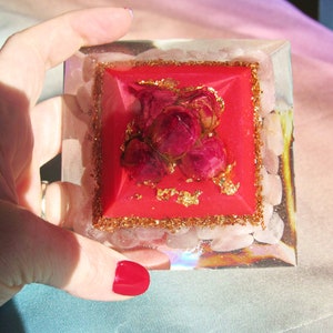 May include: A red crystal pyramid with a rose quartz base and a gold glitter border. The pyramid is topped with a cluster of dried red roses.