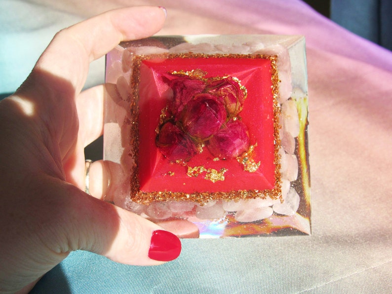 May include: A red crystal pyramid with a rose quartz base and a gold glitter border. The pyramid is topped with a cluster of dried red roses.