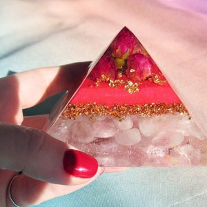 May include: A clear glass pyramid with layers of pink and gold glitter, rose quartz crystals, and dried pink roses.