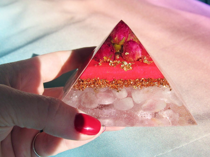 May include: A clear glass pyramid with layers of pink and gold glitter, rose quartz crystals, and dried pink roses.