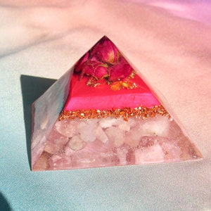 May include: A clear pink crystal pyramid with a layer of pink resin and dried pink roses at the top. The pyramid is sitting on a light blue and pink fabric.