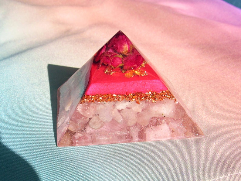 May include: A clear pink crystal pyramid with a layer of pink resin and dried pink roses at the top. The pyramid is sitting on a light blue and pink fabric.