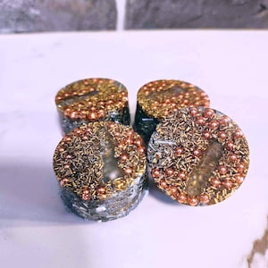 Puede incluir: Cuatro posavasos redondos de color gris, hechos de resina, con cuentas doradas y cobrizas y purpurina incrustadas en la resina. Los posavasos están dispuestos en forma de semicírculo.