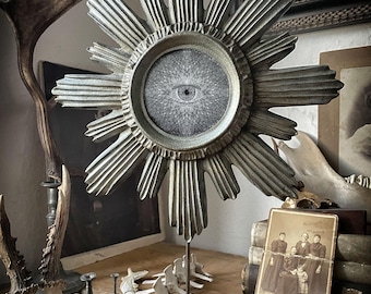 Pode incluir: Uma escultura em forma de sol prateado com um medalhão circular central apresentando uma ilustração detalhada em preto e branco de um olho. A escultura é montada numa haste de metal fina fixada a uma base quadrada de metal enferrujado.
