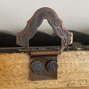 May include: A copper-toned metal D-ring hanger with an embossed leaf pattern attached to a light-colored wooden surface. The hanger is secured by two dark, circular metal fasteners, showing a weathered finish on the wood frame edge.