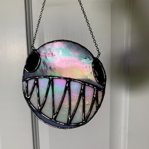 Könnte beinhalten: Ein runder, schillernder Glas-Suncatcher mit einem lächelnden Gesichtsdesign. Das Stück hat schwarze Augen, ein breites, zahnreiches Grinsen und wird von einer dunklen Metallkette gehalten. Das Glas reflektiert ein Farbspektrum, darunter Rosa, Blau und Grün.