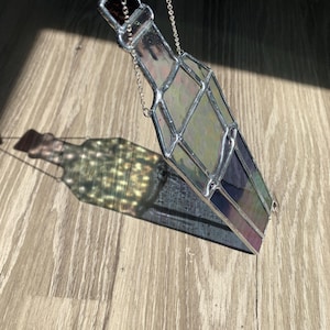 May include: A stained glass bottle-shaped suncatcher with a silver chain. The glass has a mix of colors, including purple, green, and clear. The suncatcher is hanging in front of a wooden surface, casting a colorful shadow.