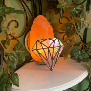 Puede incluir: Una lámpara de cristal de colores en forma de diamante con colores iridiscentes se encuentra sobre una repisa blanca junto a una lámpara de sal del Himalaya. La repisa forma parte de una estantería de esquina de metal negro con una planta verde de hojas que crece en ella.