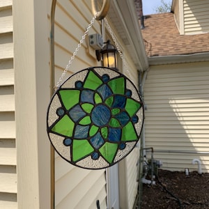 Puede incluir: Una ventana redonda de vidrieras colgando de un gancho dorado. La ventana presenta un diseño simétrico con piezas de vidrio verdes y azules. El vidrio está sujeto por líneas de metal oscuro, creando un atrapasol decorativo.