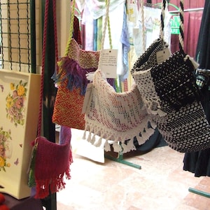 K&ouml;nnte beinhalten: Sortiment handgefertigter H&auml;keltaschen in verschiedenen Farben und Mustern, darunter Rot, Wei&szlig;, Schwarz und Blau. Die Taschen werden an einem St&auml;nder auf einem Markt ausgestellt.