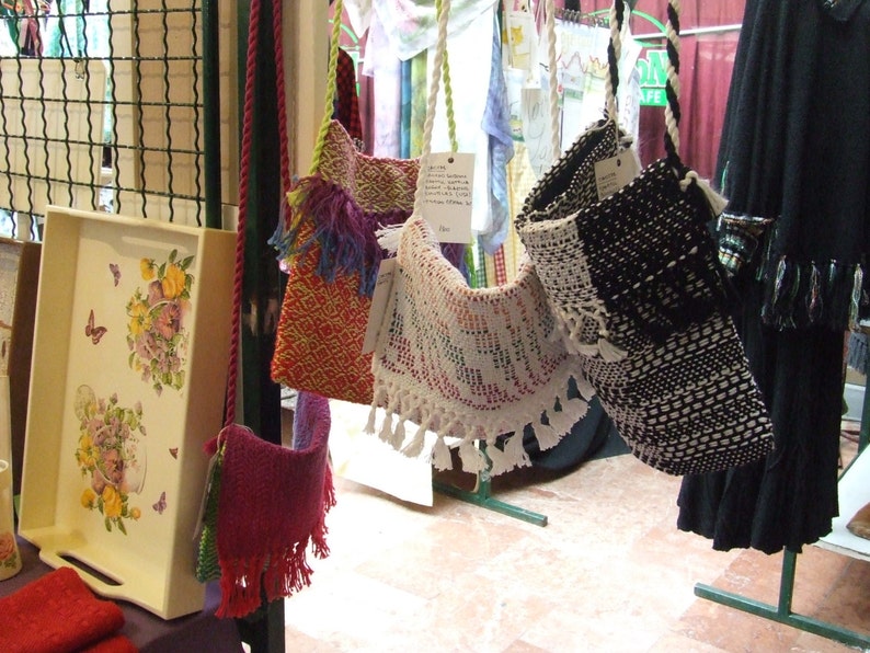 K&ouml;nnte beinhalten: Sortiment handgefertigter H&auml;keltaschen in verschiedenen Farben und Mustern, darunter Rot, Wei&szlig;, Schwarz und Blau. Die Taschen werden an einem St&auml;nder auf einem Markt ausgestellt.