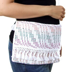 K&ouml;nnte beinhalten: Eine wei&szlig;e geh&auml;kelte Umh&auml;ngetasche mit Fransenbesatz und einem bunten geometrischen Muster. Die Tasche wird von einer Person mit einem schwarzen Oberteil und einer blauen Jeans getragen.