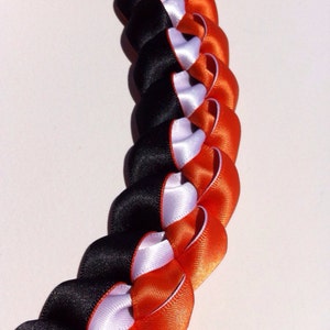 May include: A black, white, and orange braided ribbon lei. The lei is made of three different colors of ribbon that are braided together.