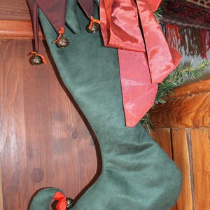 May include: A green Christmas stocking with a curled toe and a burgundy top with bells. A red ribbon and bow are attached to the stocking. The stocking is hanging on a wooden door.