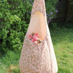 May include: A pink fabric hanging toy storage bag with a floral pattern, suspended from a green cord. The bag is filled with pink and brown balls. The background is a green lawn and foliage.