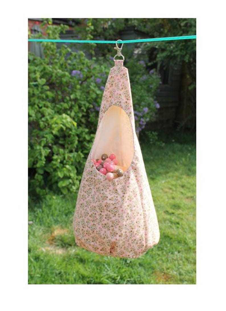 May include: A pink fabric hanging bird feeder with a floral pattern and a large oval opening. The feeder is filled with small, colorful balls and is suspended from a green line with a silver clasp.