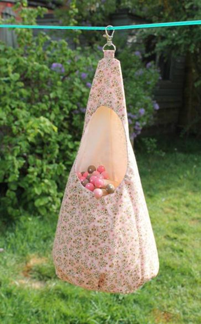 May include: A pink fabric hanging toy storage bag with a floral pattern, suspended from a green cord. The bag is filled with pink and brown balls. The background is a green lawn and foliage.