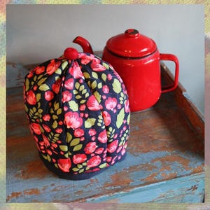 Puede incluir: Un calentador de té floral con un fondo azul marino y flores rosas y rojas, junto a una tetera roja esmaltada. El calentador tiene forma redondeada y una pequeña bola roja en la parte superior. La tetera tiene un asa y una tapa negras.