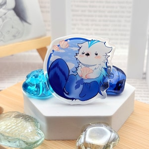 Puede incluir: Un amuleto de acrílico transparente con una nutria de dibujos animados con pelaje azul y blanco. La nutria sostiene una concha y está rodeada de agua azul y burbujas blancas.