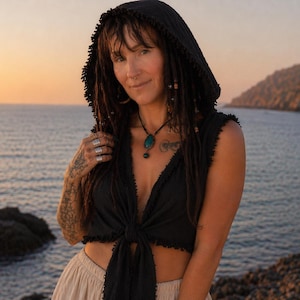 May include: A black hooded top with a plunging neckline, tied at the waist. The top is sleeveless and has a fringed edge. The person is wearing a necklace with a turquoise pendant. The background is a seascape with a sunset.