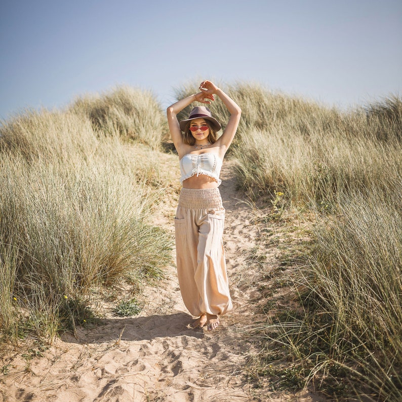 May include: A woman wearing a white crop top, beige pants, a brown hat, and pink sunglasses walks on a path through tall grass.