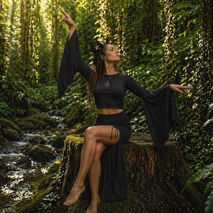 Könnte beinhalten: Ein schwarzes Crop-Top mit langen, ausgestellten Ärmeln und einem passenden Rock. Das Outfit wird in einer üppigen, grünen Waldumgebung getragen. Das Model sitzt auf einem Baumstumpf, die Arme ausgestreckt.