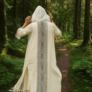 May include: A cream-colored hooded robe with a detailed black and white geometric design down the back. The robe has fringe at the sleeves and hem. The person is walking on a path through a lush green forest.