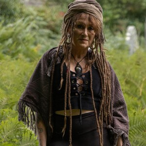 May include: A person with long dreadlocks and a headscarf, wearing a black top with a lace-up front and a fringed shawl. A necklace with a large pendant is also visible. The background is a green forest.