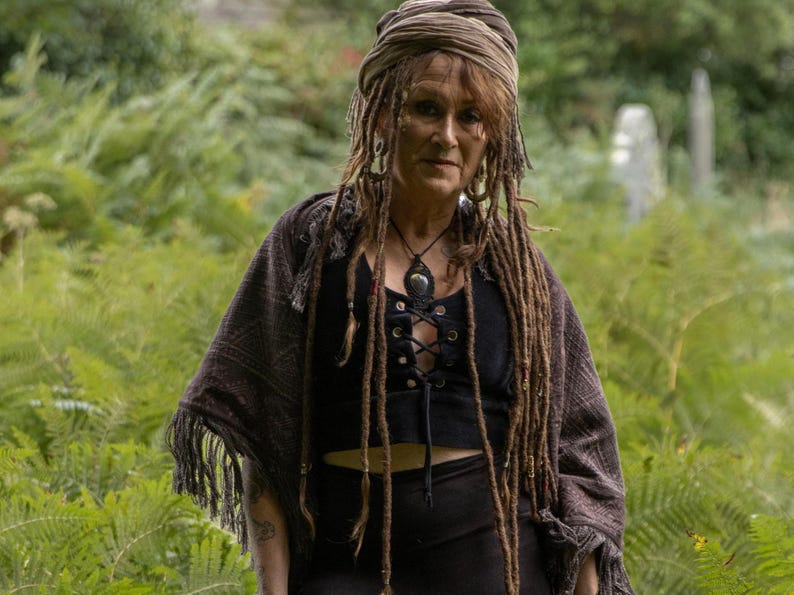 May include: A person with long dreadlocks and a headscarf, wearing a black top with a lace-up front and a fringed shawl. A necklace with a large pendant is also visible. The background is a green forest.