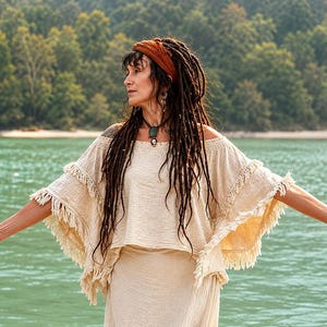 May include: A beige off-the-shoulder top featuring a wide, layered design with fringe trim along the sleeves and hem. The garment is crafted from a textured fabric and has a loose, flowing silhouette that extends to the elbows with tiered detailing.