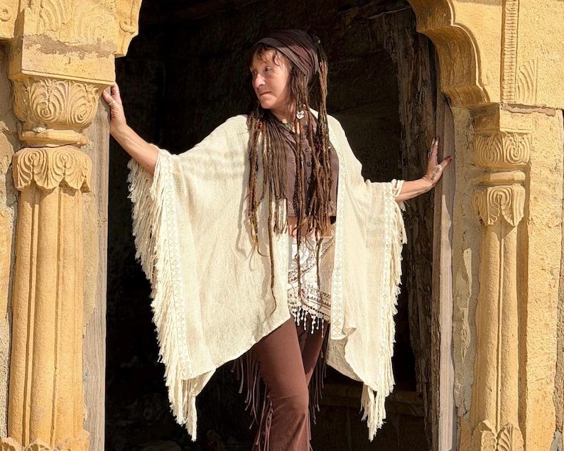 K&ouml;nnte beinhalten: Eine Frau tr&auml;gt einen cremefarbenen Poncho mit Fransen &uuml;ber einem braunen Oberteil und einer Hose. Der Poncho hat ein strukturiertes Gewebe und dekorative Fransen. Die Frau hat lange Dreadlocks und steht in einem steinernen Torbogen.