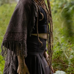 May include: A dark brown shawl with a geometric pattern and fringe detail. The shawl is draped over a black top and skirt. The image is set against a backdrop of green foliage.